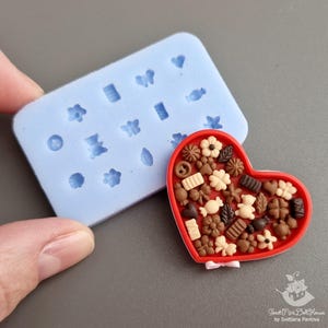 May include: A blue silicone mold with 16 different shapes for making miniature candies or decorations. The mold is next to a red heart-shaped box filled with miniature candies.