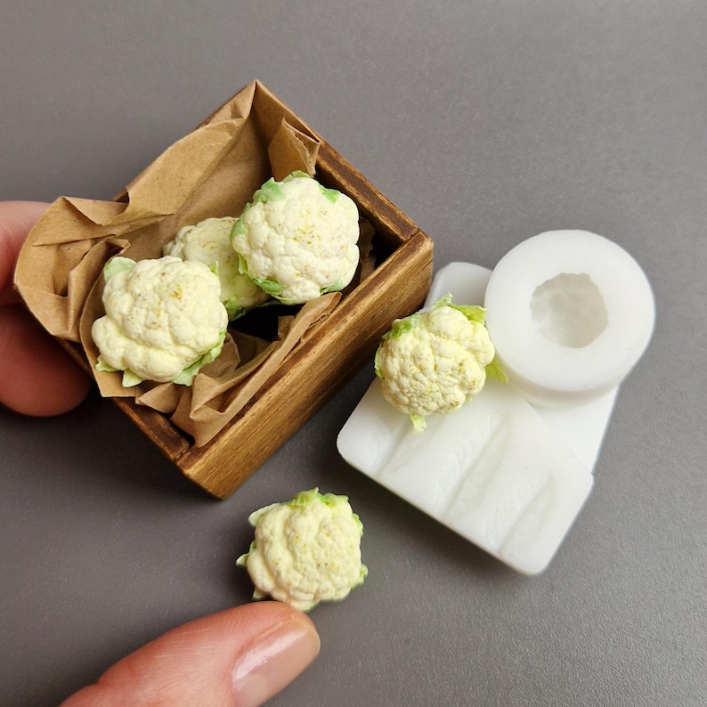 Pu&ograve; includere: Miniature di cimette di cavolfiore in una piccola scatola di legno con rivestimento di carta marrone. Diverse cimette sono anche posizionate accanto a uno stampo bianco. Il cavolfiore &egrave; bianco sporco con accenti verdi, suggerendo un accessorio artigianale o per casa delle bambole a tema alimentare.