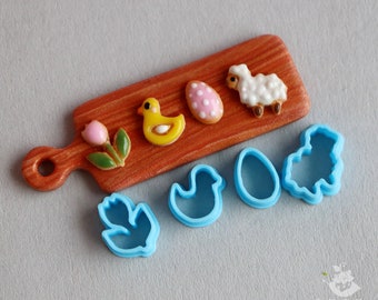 May include: A wooden cutting board with a pink flower, a yellow chick, a white lamb, and a pink and white polka dot egg cookie cutter. There are also blue plastic cookie cutters shaped like a chick, an egg, and a leaf.