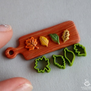 Puede incluir: Un juego de cortadores de galletas con forma de hoja en una tabla de cortar de madera. Los cortadores son verdes y la tabla es marrón. También hay tres galletas con forma de hoja de colores en la tabla.