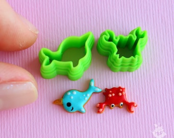 May include: Green plastic cookie cutters shaped like a narwhal and a crab, and two decorated sugar cookies, one blue narwhal and one red crab.