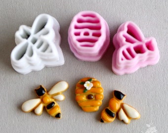 Può includere: Un set di quattro stampini per biscotti a forma di api e di un favo. Gli stampini sono in plastica rosa e bianca. Ci sono anche due biscotti decorati, uno con un'ape e uno con un favo.
