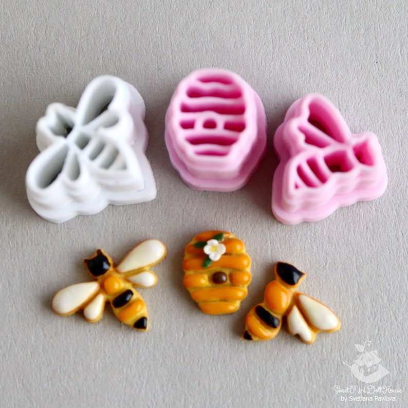 May include: A set of four cookie cutters in the shape of bees and a honeycomb. The cutters are pink and white plastic. There are also two decorated cookies, one of a bee and one of a honeycomb.