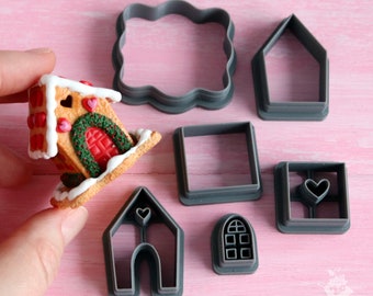 Op de afbeelding: Een set van negen grijze plastic koekjesvormen in de vorm van een gingerbreadhuis, ramen en deuren. De vormen liggen op een roze oppervlak.