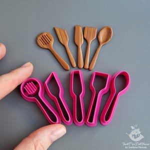 Pode incluir: Miniaturas de utensílios de cozinha de madeira e cortadores de biscoitos de plástico rosa. O conjunto de madeira inclui uma colher vazada, uma espátula e uma colher. Os cortadores rosa têm a forma dos utensílios de madeira.