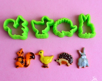 May include: Green plastic cookie cutters shaped like a squirrel, duck, hedgehog, and rabbit.  Next to the cutters are decorated cookies in the same shapes.