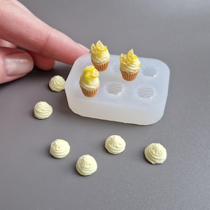 May include: Miniature lemon cupcakes with yellow icing and lemon slices, plus small dollops of white icing. The cupcakes are in a silicone mould. The items are on a grey surface.