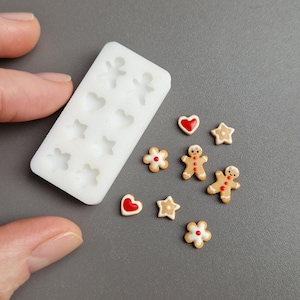Può includere: Uno stampo in silicone bianco con varie forme, tra cui cuori, stelle e omini di pan di zenzero. Diversi omini di pan di zenzero in miniatura, cuori e stelle con accenti rossi e bianchi sono nelle vicinanze.