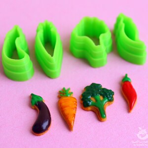 May include: Green plastic cookie cutters shaped like a carrot, eggplant, broccoli, and chili pepper.  Next to the cutters are four cookies decorated to look like the vegetables.