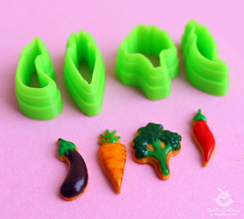 May include: Green plastic cookie cutters shaped like a carrot, eggplant, broccoli, and chili pepper.  Next to the cutters are four cookies decorated to look like the vegetables.