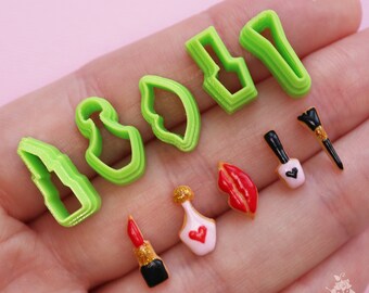 May include: A set of green plastic cookie cutters shaped like lipstick, perfume bottles, nail polish, and lips. The cutters are arranged on a hand with several decorated cookies in the background.