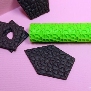 Puede incluir: Un rodillo de silicona verde con una superficie texturizada, utilizado para crear un patr&oacute;n de piedra en fondant u otros materiales comestibles. Tambi&eacute;n se muestran tres piezas de fondant negras con el mismo patr&oacute;n de piedra.