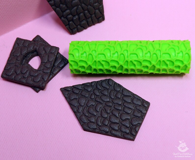 Puede incluir: Un rodillo de silicona verde con una superficie texturizada, utilizado para crear un patr&oacute;n de piedra en fondant u otros materiales comestibles. Tambi&eacute;n se muestran tres piezas de fondant negras con el mismo patr&oacute;n de piedra.