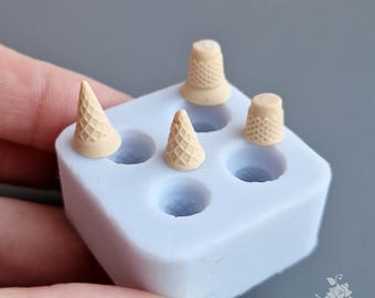 Puede incluir: Un molde de silicona azul claro con cuatro cavidades, que contiene mini conos de helado y dedales beige. Los artículos están dispuestos en el molde, con dos conos y dos dedales. El molde es sostenido por una mano sobre un fondo gris.