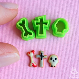 Puede incluir: Cortadores de galletas de plástico verde en forma de cruz, hueso y calavera. También hay tres galletas pequeñas decoradas: un hueso, una cruz y una calavera.