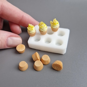 以下が含まれることがあります： 黄色いアイシングとレモンスライスを添えたミニチュアのレモンカップケーキ。白い型と個別のカップケーキライナーの隣に配置されています。これらは、ドールハウスやクラフトプロジェクトに最適な手作りの食品アイテムです。