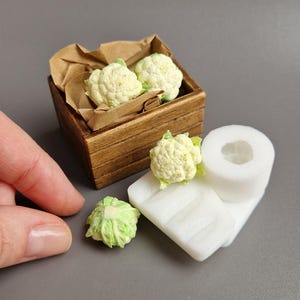 Könnte beinhalten: Miniatur-Blumenkohlköpfe, einige in einer Holzkiste mit braunem Papier, andere auf einer weißen Oberfläche. Ein Blumenkohlkopf ist separat. Die Miniaturgemüse sind cremefarben mit grünen Akzenten, perfekt für Puppenhaus-Essen oder Bastelprojekte.
