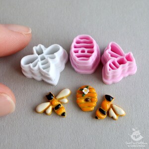 May include: A set of bee-themed cookie cutters, including a honeycomb, a bee, and a beehive. The cutters are made of white and pink plastic. There are also two small, decorated cookies in the shape of a bee and a honeycomb.
