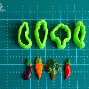 May include: Set of six green plastic cookie cutters shaped like vegetables, including a carrot, eggplant, pepper, broccoli, and two different types of leaves.  The cutters are arranged on a green cutting mat with white dots.