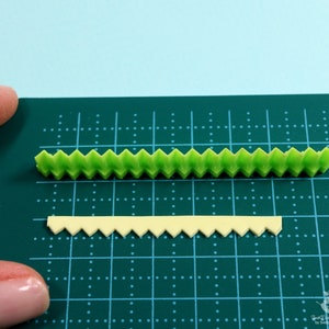 Może przedstawiać: Dwa paski dekoracyjnego lukru, jeden zielony, a drugi ż&oacute;łty, z falistym brzegiem. Lukier leży na zielonej macie do krojenia.