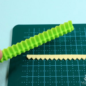 Może przedstawiać: Zielone plastikowe narzędzie z wzorem zygzakowatym, używane do tworzenia ozdobnych krawędzi na fondancie lub innych jadalnych materiałach. Na zdjęciu widoczna jest r&oacute;wnież mniejsza, ż&oacute;łta wersja narzędzia.