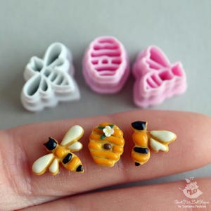 May include: A set of bee-themed cookie cutters, including a honeycomb, a bee, and a beehive. The cutters are made of white and pink plastic.