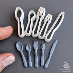 May include: Miniature dollhouse cutlery set. The set includes spoons, forks, and knives in two styles: white plastic cutters and grey, finished pieces. The pieces are arranged on a grey surface.