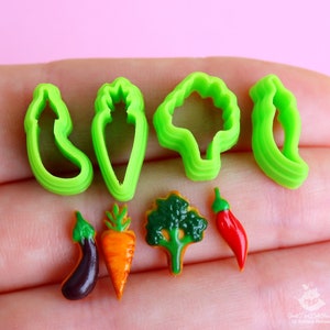 May include: A set of green plastic cookie cutters shaped like vegetables, including a carrot, broccoli, pepper, and eggplant. There are also miniature plastic vegetables, including a carrot, eggplant, broccoli, and a red chili pepper.