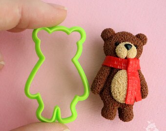 May include: Green plastic bear cookie cutter next to a brown and beige textured bear figurine wearing a red scarf.