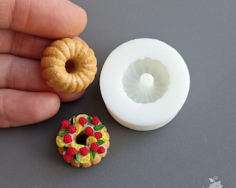 May include: A miniature, detailed polymer clay donut with a textured surface, next to a white silicone mold. A second donut is decorated with red berries and lemon slices. The donuts are on a gray surface.