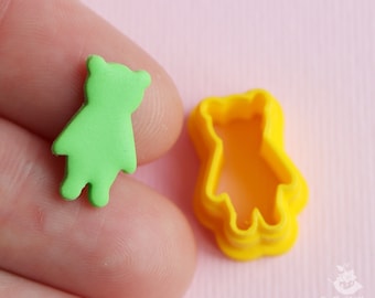 May include: A small, lime green bear-shaped polymer clay figure next to a yellow bear-shaped cookie cutter. The bear figure has a simple design with rounded features. The cookie cutter is made of yellow plastic.