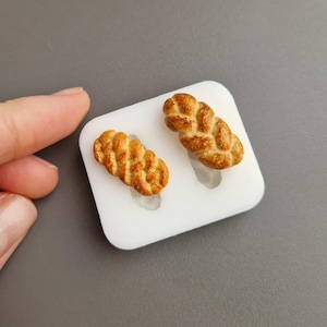 Op de afbeelding: Twee miniatuur gevlochten broodjes, met een goudbruine korst, liggen in een witte mal. Het brood heeft gedetailleerde texturen, wat duidt op een vers gebakken product. De achtergrond is effen grijs, wat een contrast vormt met de voedingsmiddelen. De afbeelding richt zich op het miniatuurvoedsel.