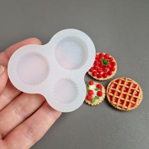 May include: A translucent silicone mould with three circular cavities of varying sizes, alongside three miniature food replicas: a round tart topped with red berries, a smaller tart with white cream and red berries, and a lattice-topped fruit pie.