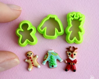 Op de afbeelding: Een set van vijf kerstthema-koekjesvormen, waaronder een peperkoekman, een rendier, een trui en twee verschillende trui-ontwerpen. De vormen zijn groen en de koekjes zijn versierd met glazuur.