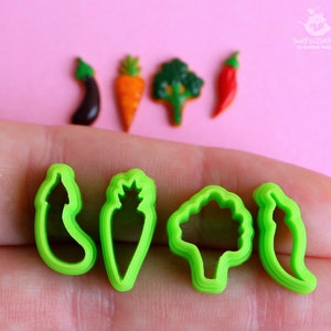 May include: Four green plastic cookie cutters shaped like a pepper, carrot, broccoli, and eggplant. The cutters are on a hand against a pink background.