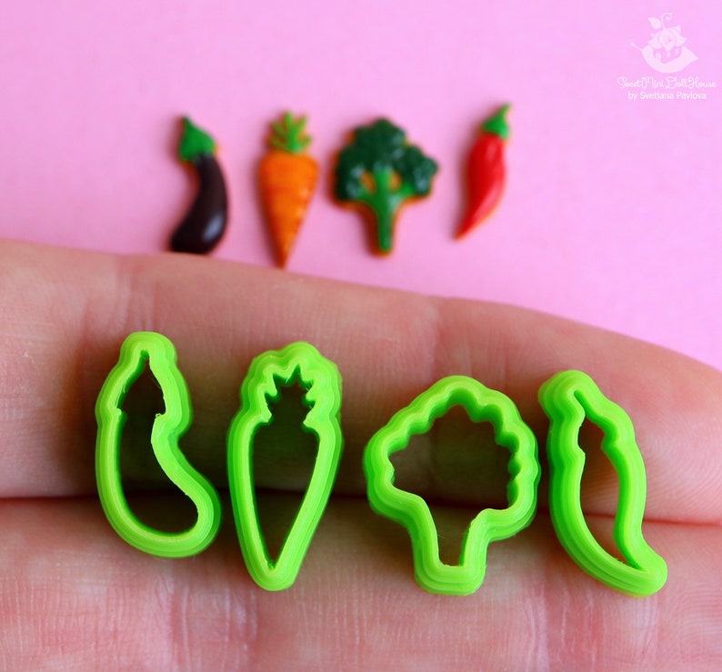 May include: Four green plastic cookie cutters shaped like a pepper, carrot, broccoli, and eggplant. The cutters are on a hand against a pink background.