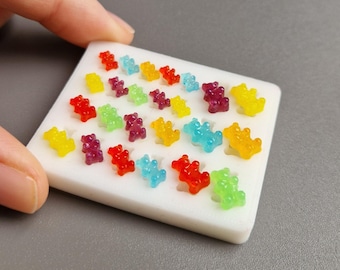 Könnte beinhalten: Eine weiße Schale gefüllt mit bunten Gummibärchen. Die Gummibärchen sind in verschiedenen Farben, darunter Rot, Gelb, Grün, Lila und Blau. Die Schale wird von einer Hand gehalten, der Hintergrund ist einfarbig grau.