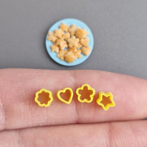 Könnte beinhalten: Miniatur-Lebensmittel. Ein hellblauer Teller enthält winzige stern-, blumen- und herzförmige Kekse. Vier gelbe Ausstecher in Form einer Blume, eines Herzens, einer Blume und eines Sterns befinden sich im Vordergrund. Der Hintergrund ist eine graue Oberfläche.