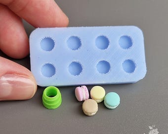 Puede incluir: Un molde de silicona azul claro con ocho cavidades circulares, junto a macarons en miniatura en rosa, amarillo, beige y azul. También hay un pequeño cilindro verde. Los artículos están sobre una superficie gris. La imagen es de comida en miniatura.