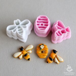 May include: A set of four cookie cutters in the shape of bees and a honeycomb. The cutters are made of pink and white plastic. There are also two decorated cookies, one a bee and the other a honeycomb.
