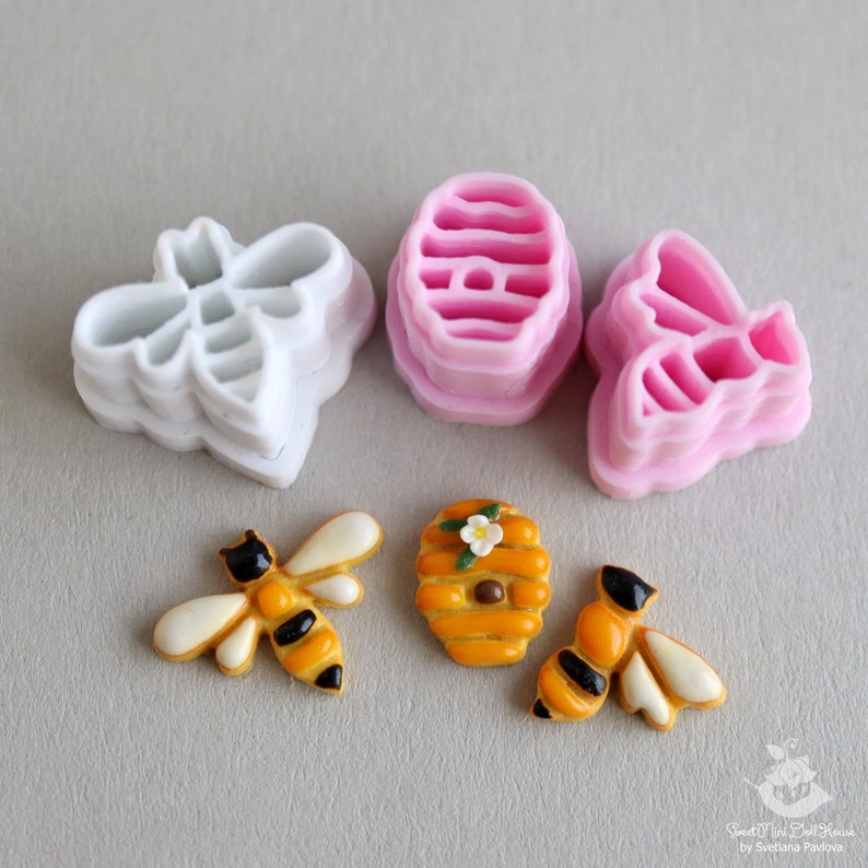 May include: A set of four cookie cutters in the shape of bees and a honeycomb. The cutters are made of pink and white plastic. There are also two decorated cookies, one a bee and the other a honeycomb.