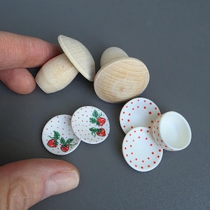 Peut inclure: Deux formes de champignons en bois clair non fini accompagnées de quatre assiettes miniatures en céramique blanche et d'un petit bol en céramique blanche, tous décorés de petits pois rouges et de motifs de fraises peints.