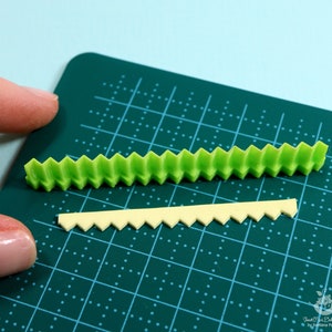 Może przedstawiać: Dwa paski ozdobnej listwy, jeden zielony, a drugi ż&oacute;łty, z falistym brzegiem. Listwa leży na zielonej macie do cięcia.