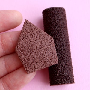 Könnte beinhalten: Zwei braune, strukturierte Objekte auf rosa Hintergrund. Eines ist rautenförmig, das andere zylindrisch. Die Objekte haben eine raue, poröse Oberfläche. Die Rautenform wird von einer Hand gehalten.