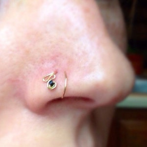 Puede incluir: Un aro de nariz de oro con una pequeña piedra preciosa verde oscuro. El aro se lleva en un piercing en la nariz.