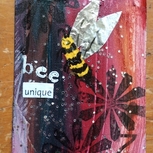 May include: Mixed media artwork featuring a bee with yellow and black stripes and silver foil wings. The background transitions from dark to red with floral and glitter accents. The words "bee unique" are displayed.