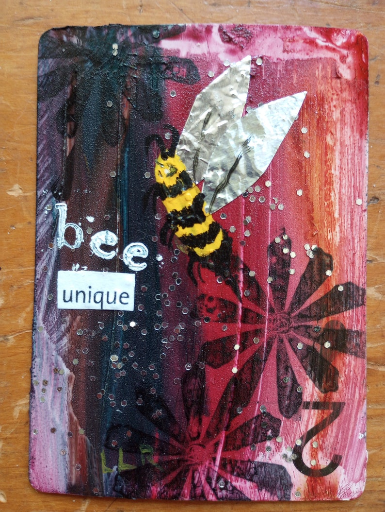 May include: Mixed media artwork featuring a bee with yellow and black stripes and silver foil wings. The background transitions from dark to red with floral and glitter accents. The words "bee unique" are displayed.