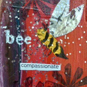 May include: A mixed media artwork featuring a yellow and black bee with silver foil wings against a red background. The word "bee" is written in white, and the word "compassionate" is below the bee. Silver glitter dots are scattered throughout.