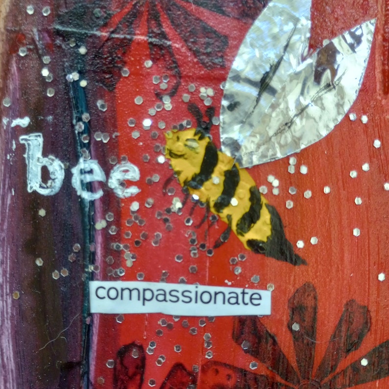 May include: A mixed media artwork featuring a yellow and black bee with silver foil wings against a red background. The word "bee" is written in white, and the word "compassionate" is below the bee. Silver glitter dots are scattered throughout.