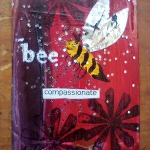 May include: A rectangular artwork with a red and purple background, featuring a yellow and black bee with silver wings. The word "bee" is written in white, and the word "compassionate" is on a white label. Glitter is scattered across the surface.